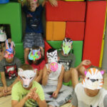 Dzieci siedzą na podłodze i na dużych klockach, na twarzach mają założone papierowe maski zwierząt.