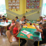 Dzieci siedzą przy stolikach i wyklejają kontury statków wydziertanką z kolorowego papieru.
