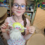 Uśmiechnięta dziewczynka w okularach prezentuje laleczkę z włóczki